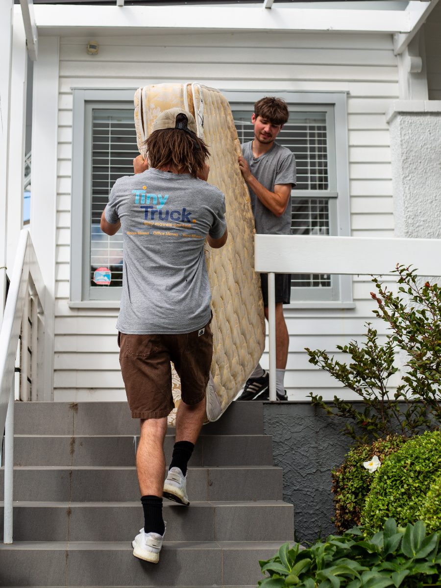 Two Tiny Truck crew carrying an old mattress down the front steps of a white weatherboard house